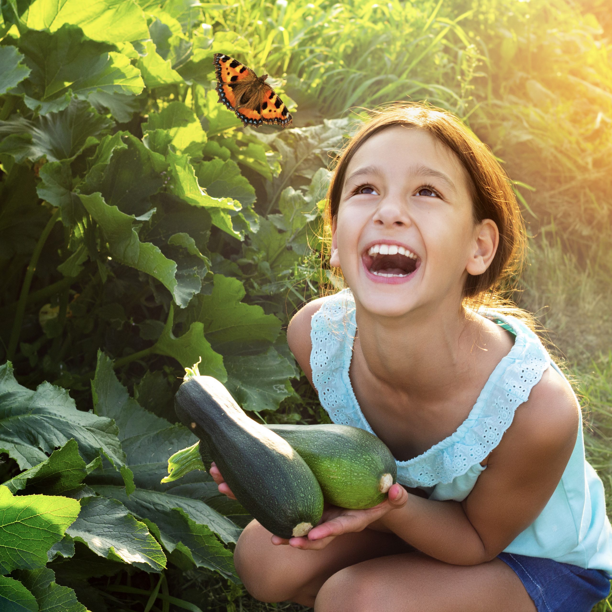 Happy laughing girl holding fresh zucchini outdoors in garden looking on flying butterfly