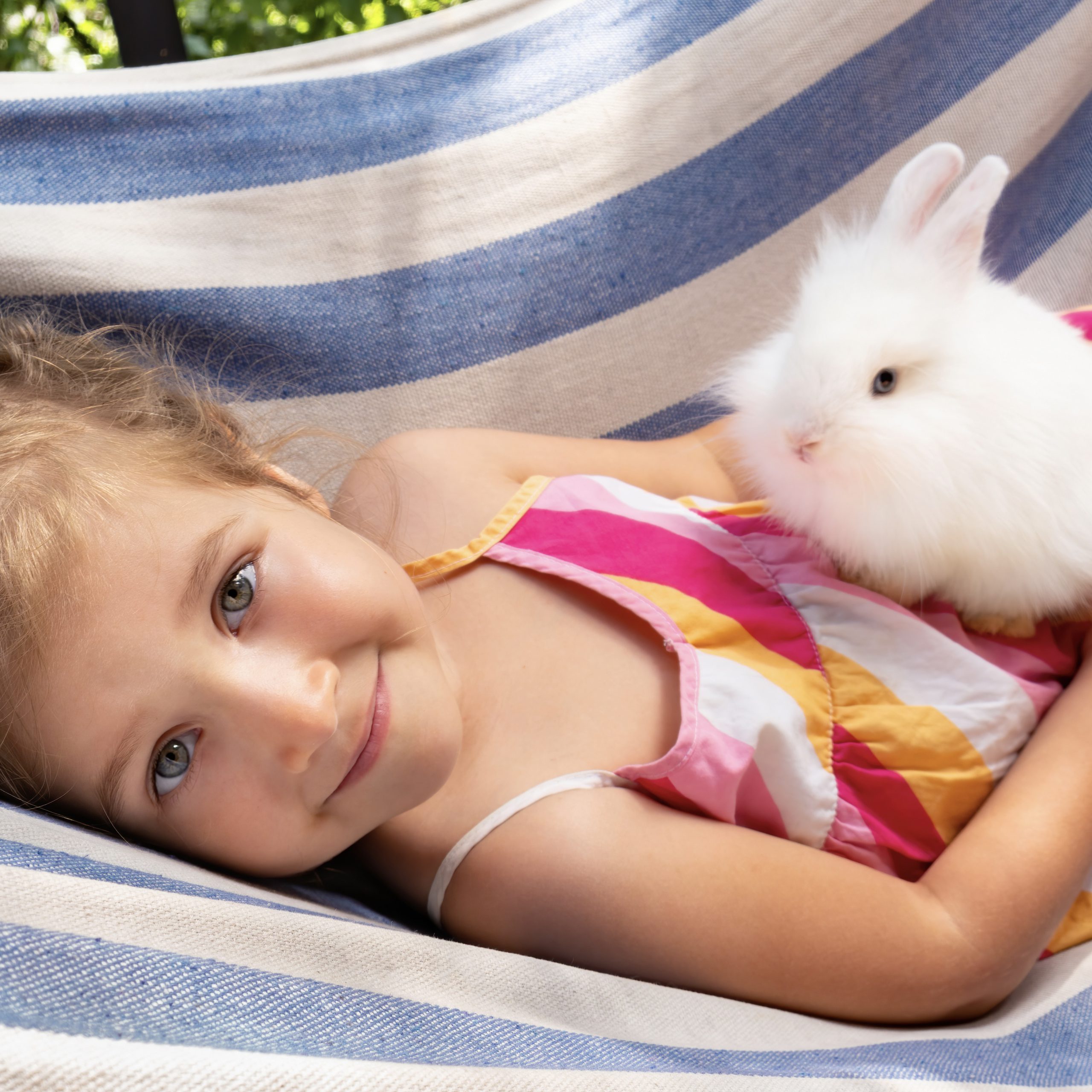 A white fluffy bunny in the arms of a cute girl. The child holds a decorative rabbit in his arms and rides on a hammock on a sunny day. Happy little girl with positive emotions smiles at her pet.